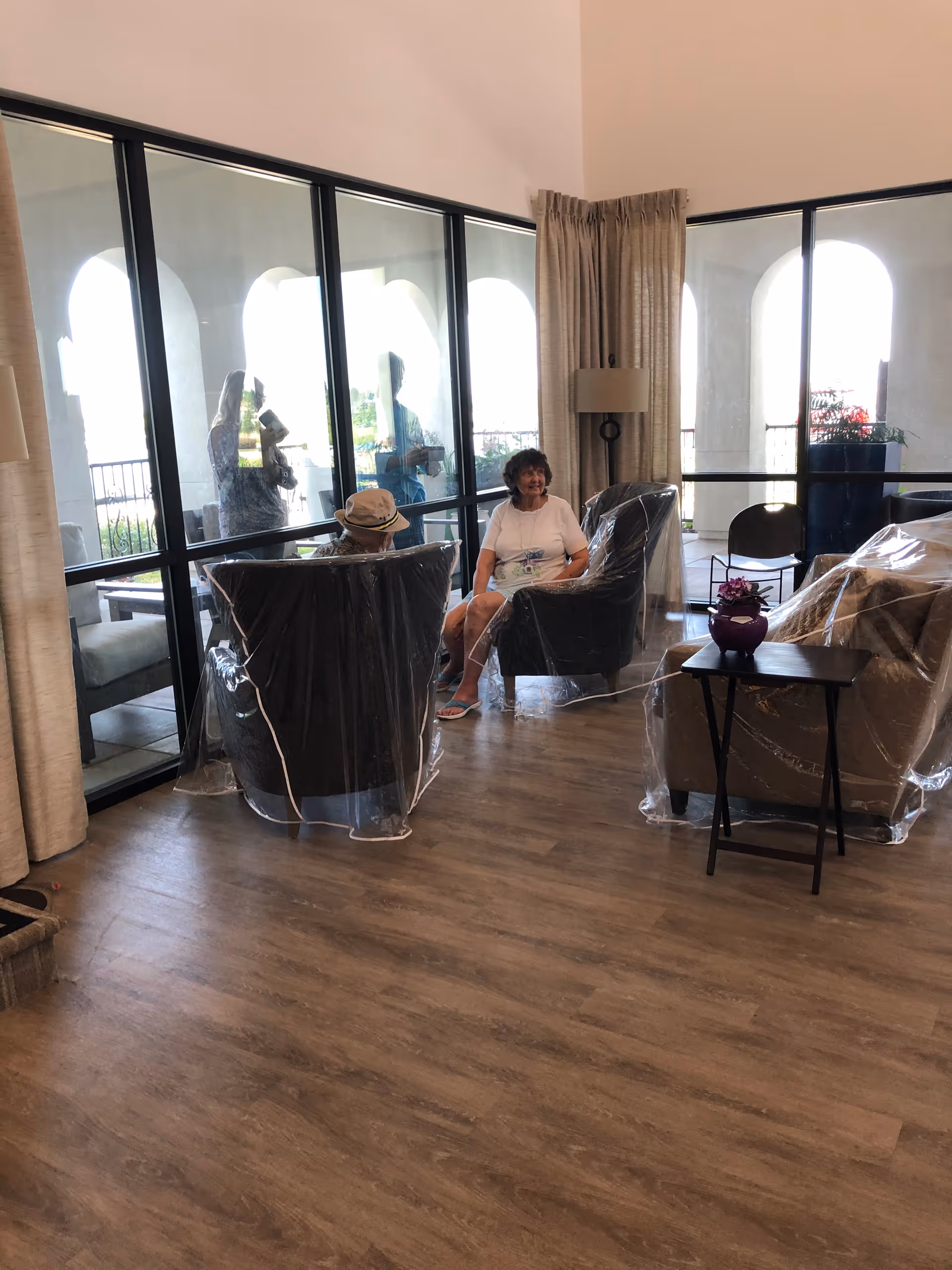A living room area with three chairs covered in protective plastic. Two elderly people are seated in two of the chairs, one wearing a hat and the other in a white shirt. Large windows with black frames and beige curtains let in natural light, showing an outdoor patio with arches. A small table with a purple vase and flower is placed near one of the chairs.