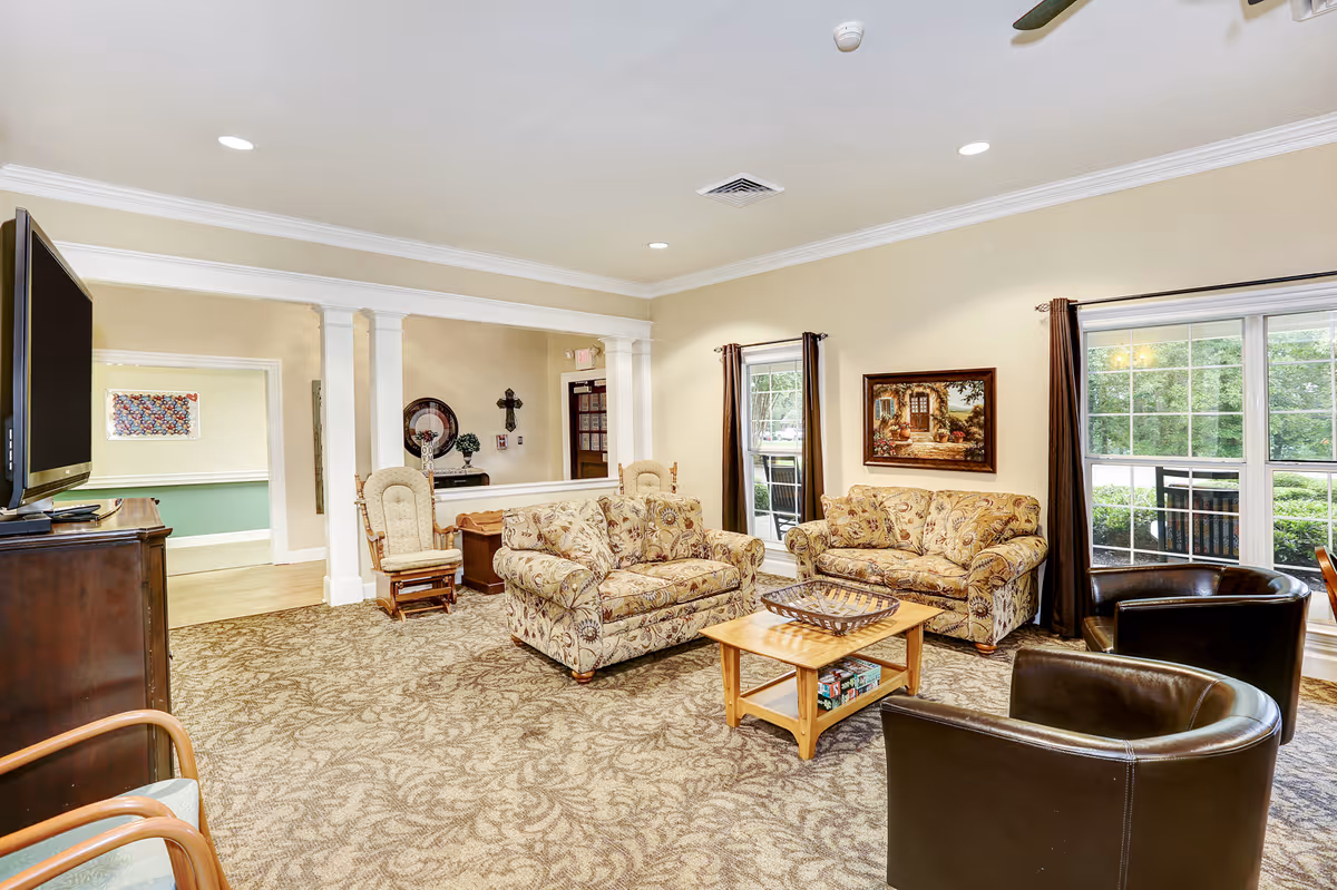 A cozy living room in Country Cottage Corinth featuring two floral patterned sofas, two dark leather armchairs, a wooden coffee table with a decorative tray, a flat-screen TV on a wooden stand, large windows with brown curtains letting in natural light, and beige walls with crown molding. The room has a patterned carpet and a ceiling fan.