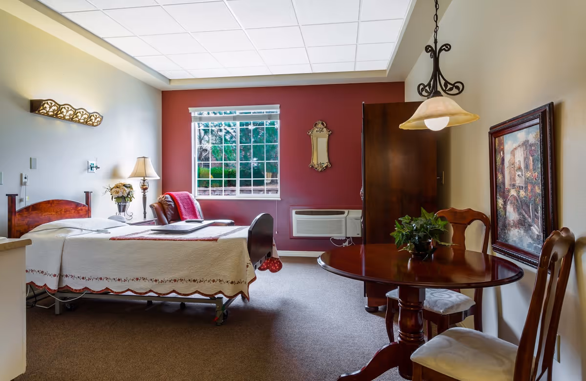 A furnished senior living bedroom with a bed, bedside lamp, window, and a small round dining table with chairs.
