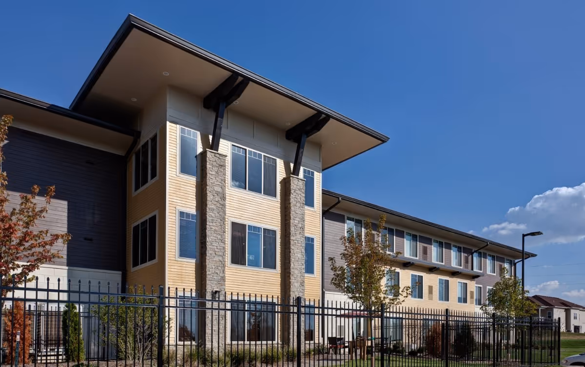 Exterior view of a modern three-story assisted living facility building with large windows, stone pillars, and a fenced garden area under a clear blue sky.