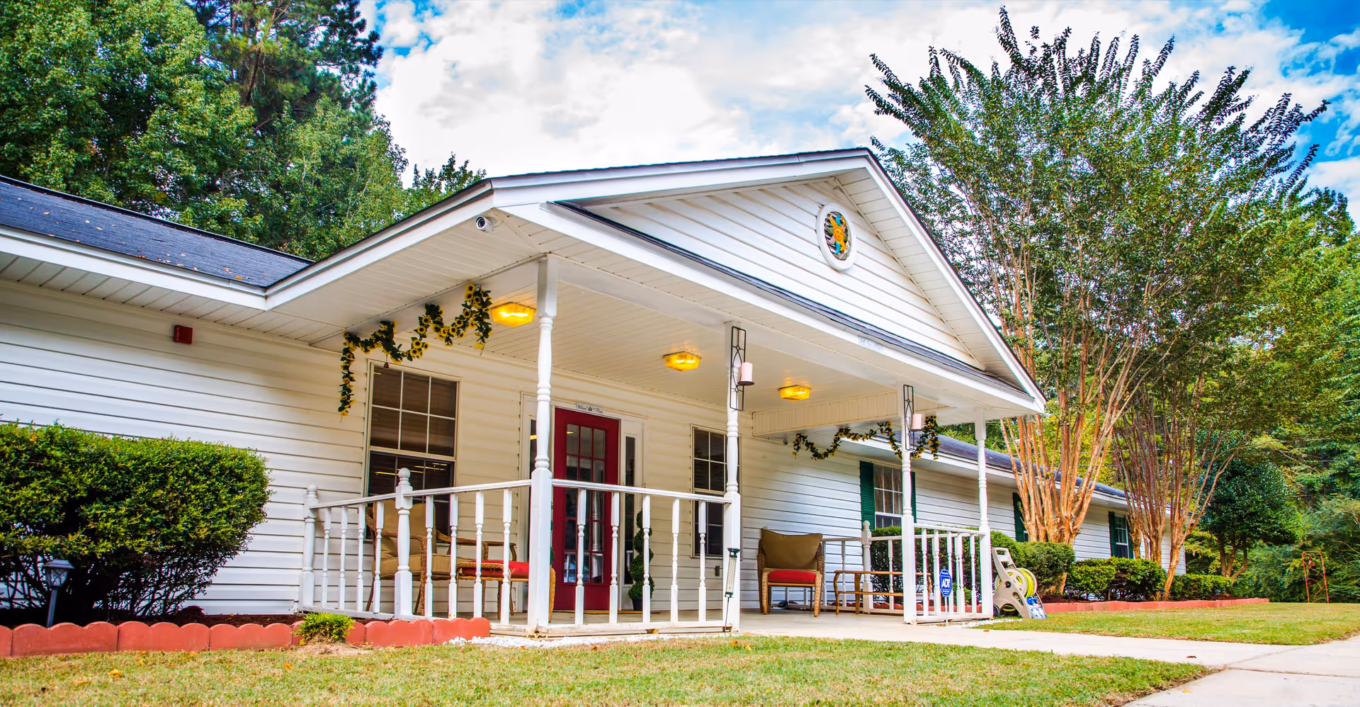 Exterior view of a single-story white senior living facility building with a covered porch decorated with garlands and outdoor seating. The building has a red door, white railings, and is surrounded by green trees and bushes under a partly cloudy sky.