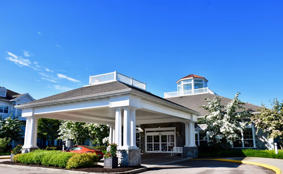 Front exterior view of StoneRidge Senior Living facility with a covered entrance supported by white columns, surrounded by greenery and flowering trees under a clear blue sky.