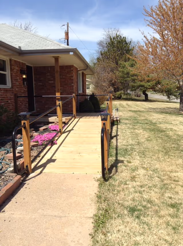 A wooden wheelchair ramp with black handrails leading to the entrance of a brick building. The ramp is surrounded by a grassy lawn with some trees and bushes, and there are pink flowers planted near the base of the ramp.