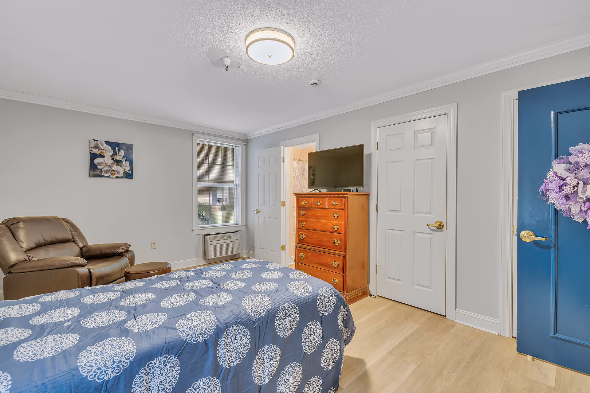 A bedroom with a bed covered in a blue and white patterned comforter, a brown leather recliner chair with a matching ottoman, a wooden dresser with a flat-screen TV on top, a window with blinds, and three doors including one painted blue with a purple wreath hanging on it.