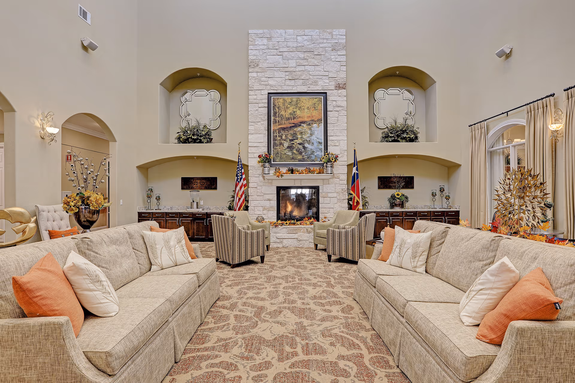A spacious and elegant living room with two beige sofas adorned with white and orange pillows facing each other across a patterned carpet. In the center background is a stone fireplace with a fire burning, flanked by two armchairs and decorated with flowers and autumn-themed decor. Above the fireplace hangs a framed painting of a serene nature scene. The walls feature built-in alcoves with decorative mirrors and plants, and there are American and Texas flags on either side of the fireplace. Large windows with beige curtains allow natural light into the room.