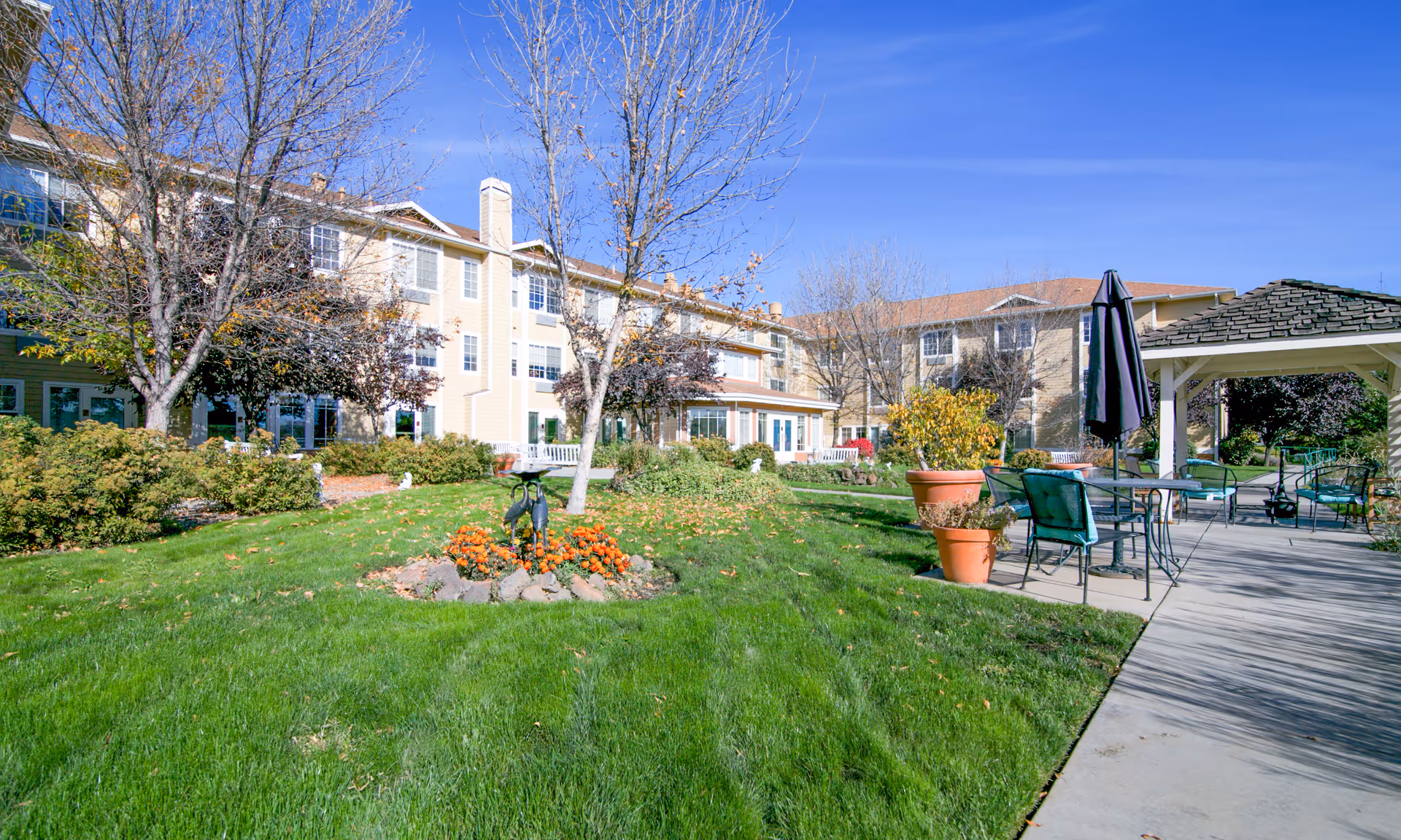 Outdoor garden area of Brookdale Yakima with green grass, leafless trees, flower beds, potted plants, patio tables and chairs with umbrellas, and a gazebo. The background shows a multi-story beige building under a clear blue sky.