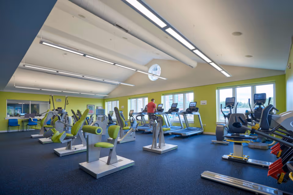 A spacious fitness room with various exercise machines including treadmills, stationary bikes, and strength training equipment. The room has large windows letting in natural light, green walls, and a high ceiling with modern lighting fixtures. A person in a red shirt is using one of the treadmills near the windows.