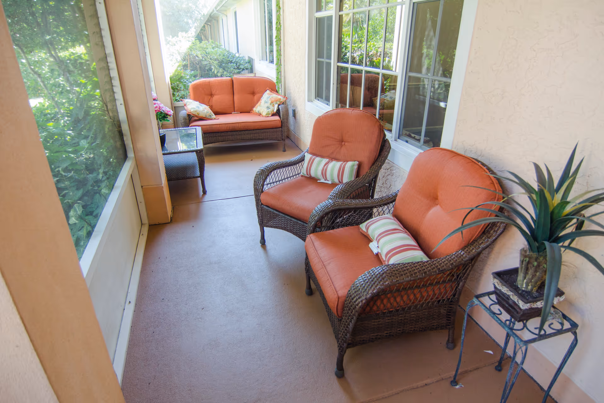 A screened-in porch area with two cushioned wicker armchairs and a cushioned wicker loveseat, all with orange cushions and striped pillows. There is a small glass-top side table between the loveseat and chairs, and a potted plant on a metal stand near the chairs. The porch overlooks greenery outside.