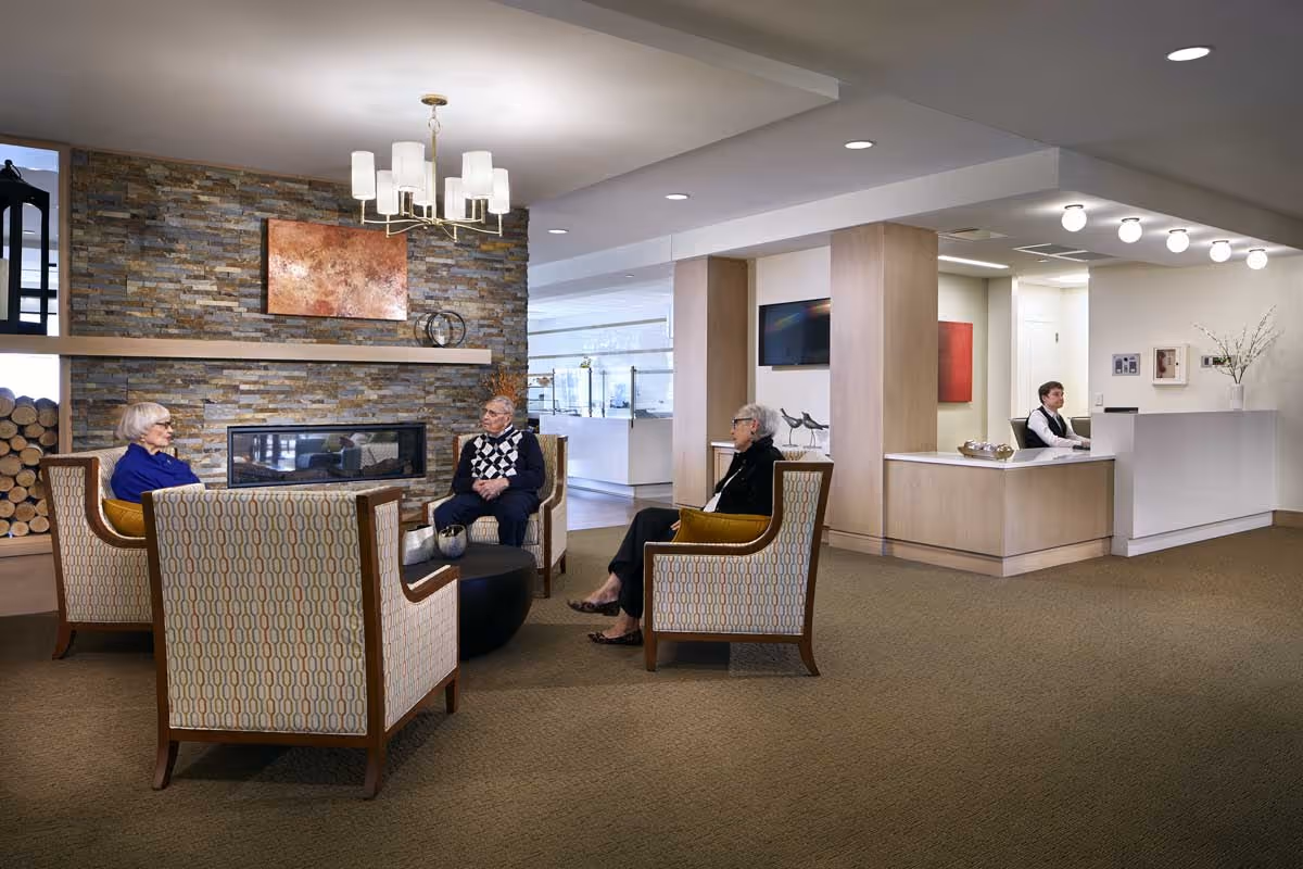 A senior living facility lobby with residents seated in armchairs around a fireplace and a receptionist at the front desk.