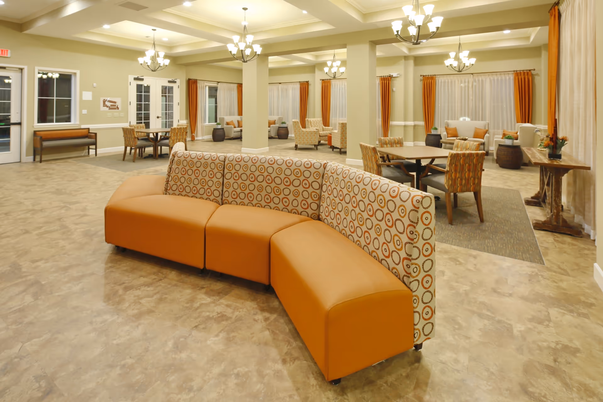 A spacious and well-lit common area in a senior living facility featuring a curved sectional sofa with orange and patterned upholstery in the foreground. The room has multiple seating areas with tables and chairs, large windows with orange curtains, and several chandeliers hanging from the ceiling. The floor is covered with a light-colored tile, and there are decorative plants and a wooden console table along the right wall.