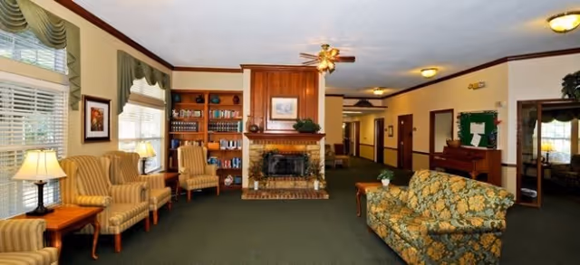 A cozy living room area with green carpet, a floral patterned sofa, two striped armchairs, wooden side tables with lamps, a brick fireplace with a wooden mantel, built-in wooden shelves with books and decor, and a ceiling fan with lights. The room has large windows with valances and a hallway leading to other rooms.