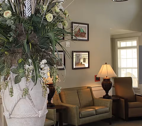 A cozy sitting area with a large floral arrangement in the foreground, upholstered sofa and armchairs, table lamps, framed wall art, and a bright window.