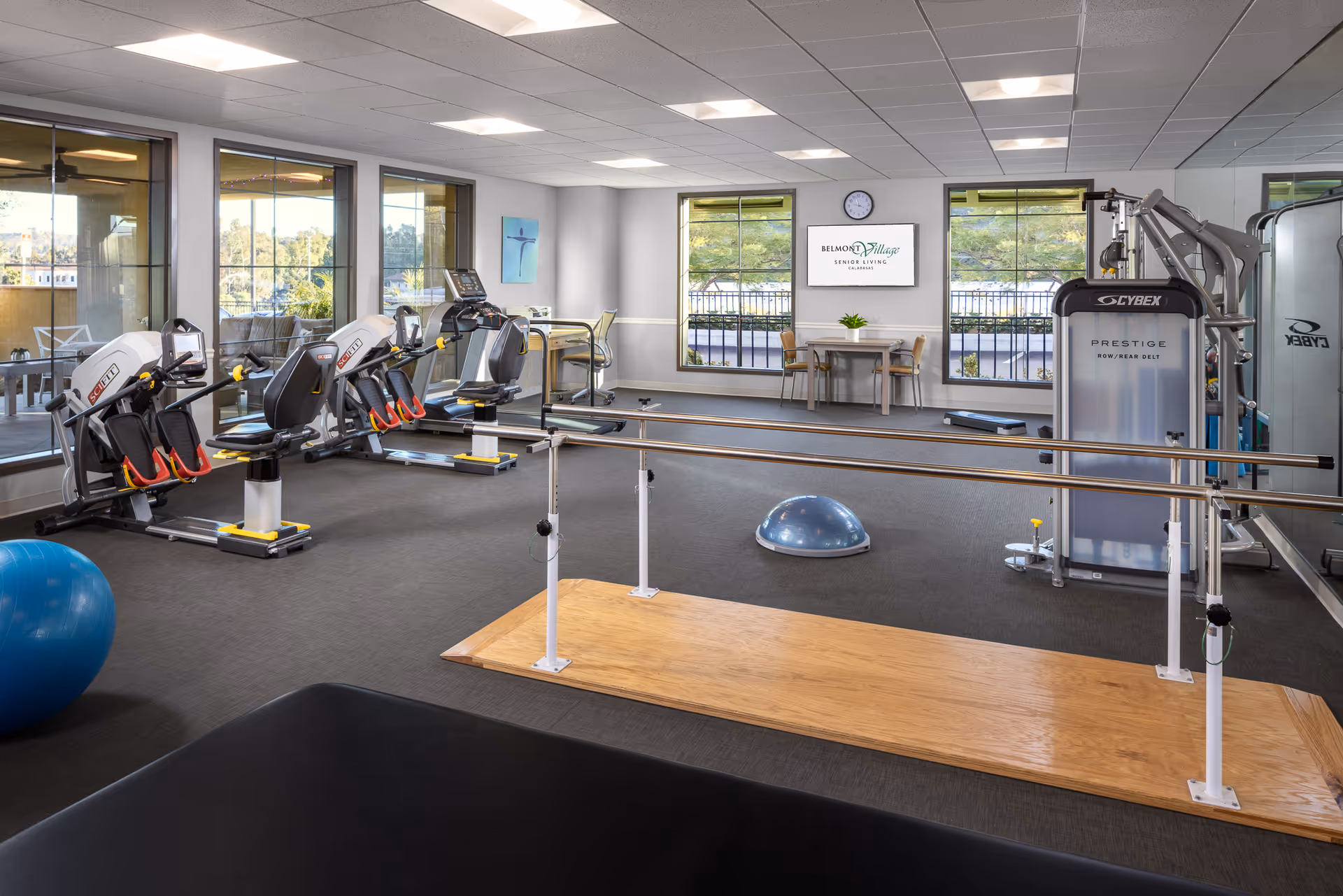 A senior living facility fitness room with exercise equipment including recumbent bikes, a multi-function strength machine, balance bars, a blue exercise ball, and a half-dome balance trainer. The room has large windows with views of greenery outside, a wall clock, and a table with chairs beneath a screen displaying the Belmont Village Senior Living Calabasas logo.
