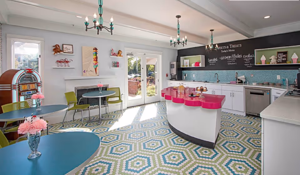 Bright retro-style communal dining and kitchenette with teal tables, patterned hexagonal tile floor, a pink-topped island, and a chalkboard menu.