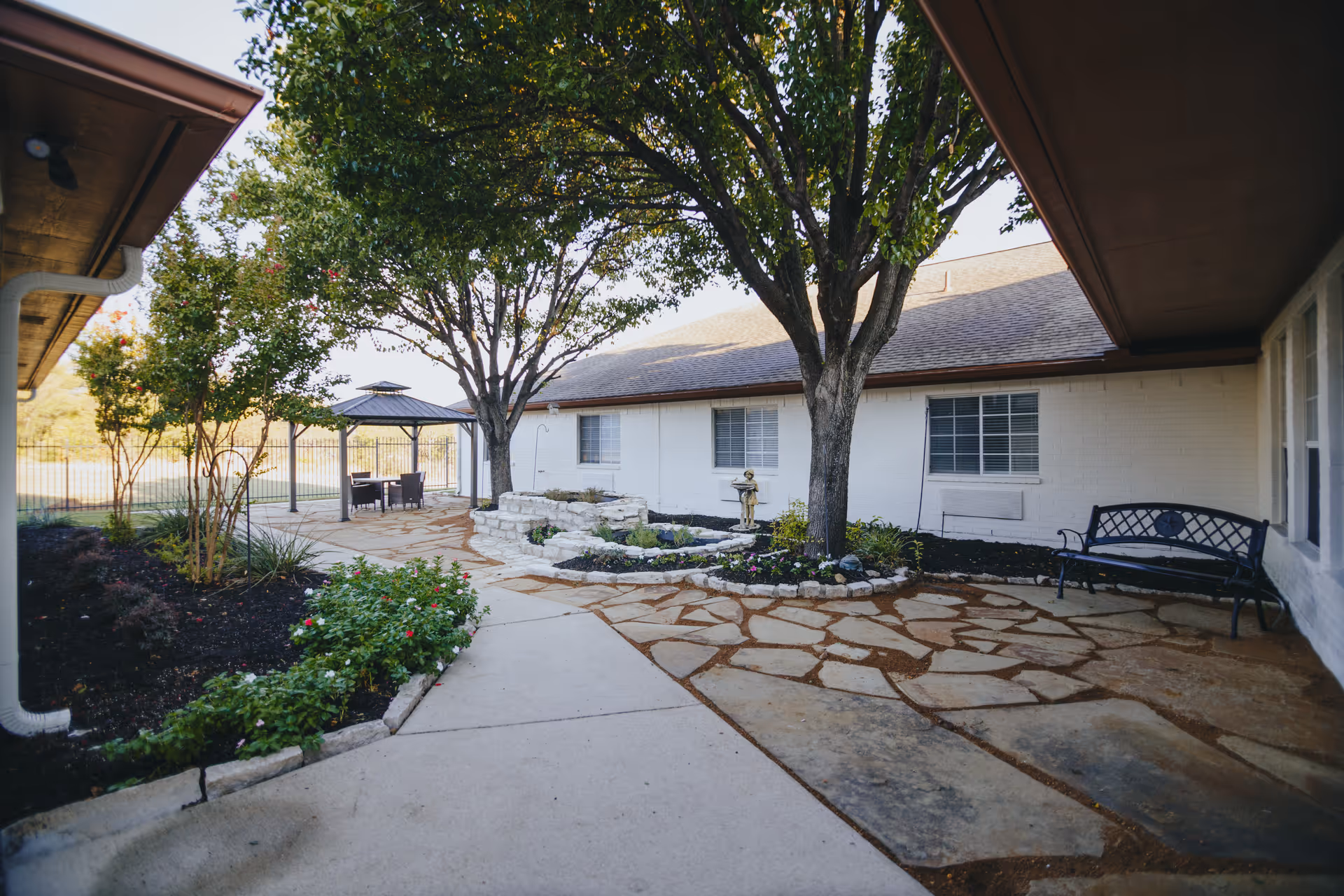 Outdoor courtyard area with a stone and concrete pathway, trees, flower beds, a small water fountain, a black metal bench, and a gazebo with a table and chairs, adjacent to a white brick building.