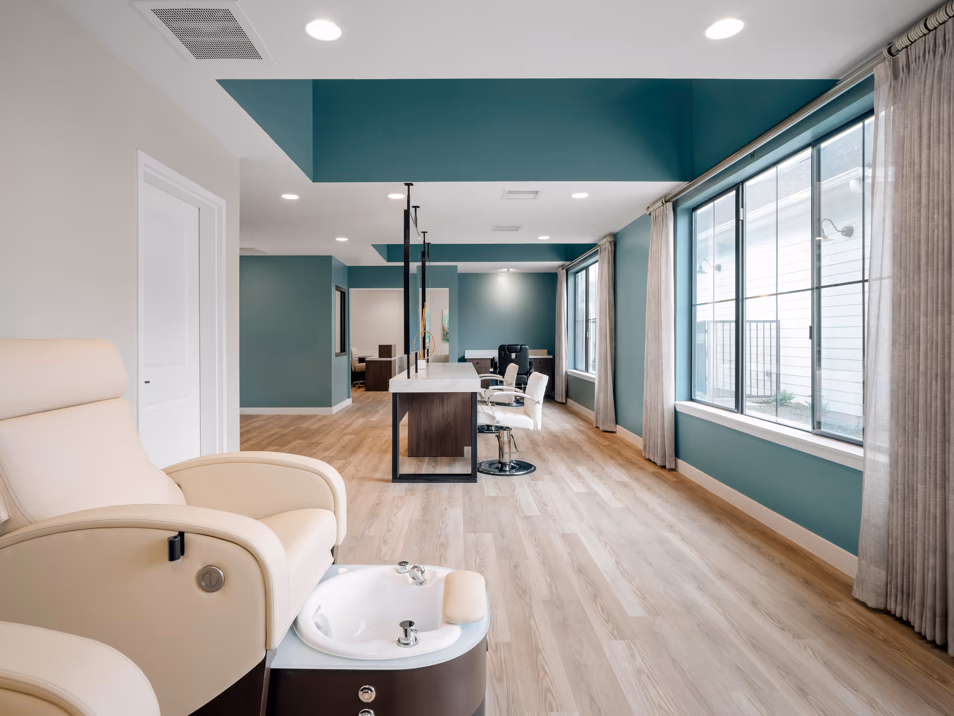 Bright interior salon area in a senior living facility with a pedicure chair in the foreground, a central manicure island, teal walls, and large windows.