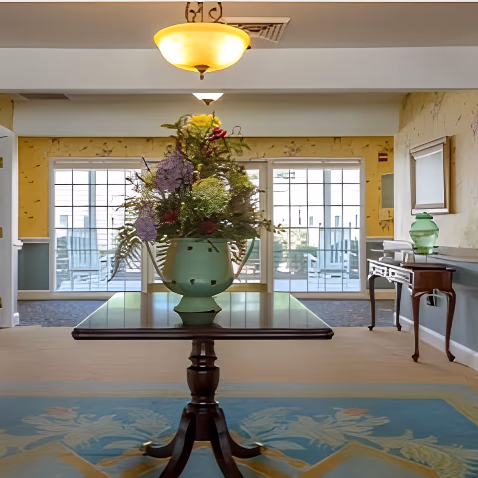 Lobby interior with a central wooden table holding a large floral arrangement and windows with seating visible in the background.