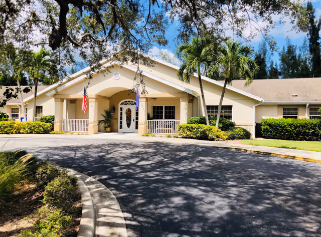 Front exterior view of Arden Courts - ProMedica Memory Care Community in Ft. Myers, showing a single-story building with a covered entrance, two flags, palm trees, and surrounding greenery under a partly cloudy sky.
