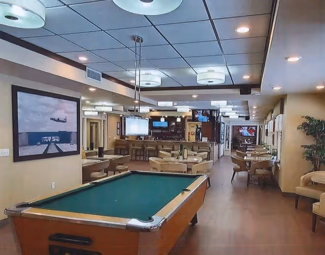 Interior view of a recreational lounge area featuring a pool table in the foreground, multiple round tables with chairs, a bar area with stools, and several mounted televisions. The room has a drop ceiling with recessed lighting and pendant lights, beige walls, and a large framed picture on the left wall.