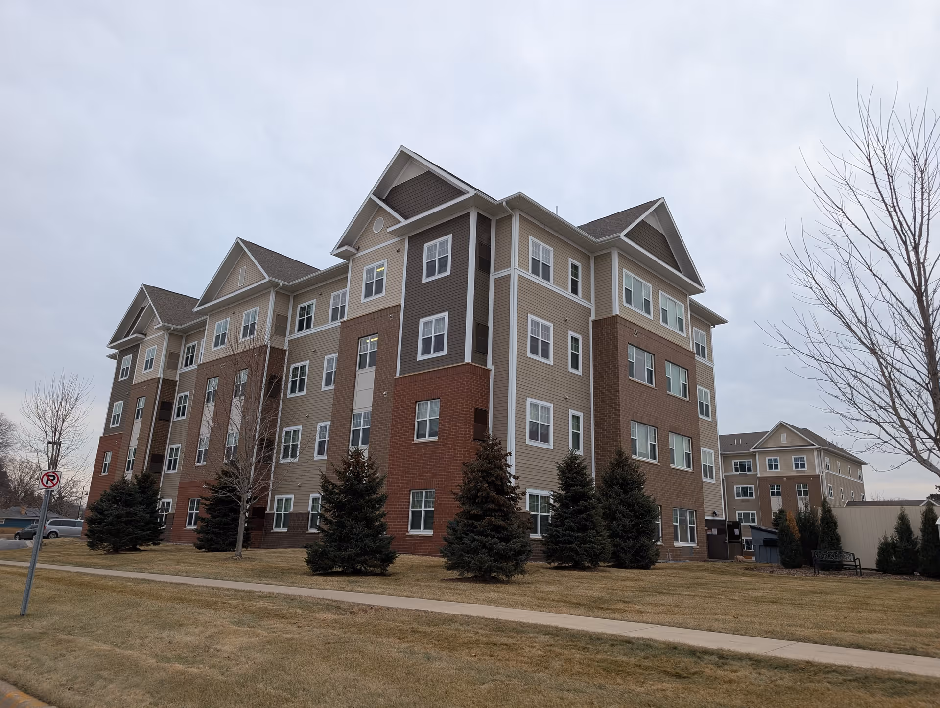 Exterior view of a multi-story senior living facility building with beige, brown, and red brick siding, multiple windows, and a sloped roof. The building is surrounded by a lawn with small evergreen trees and leafless deciduous trees under an overcast sky.