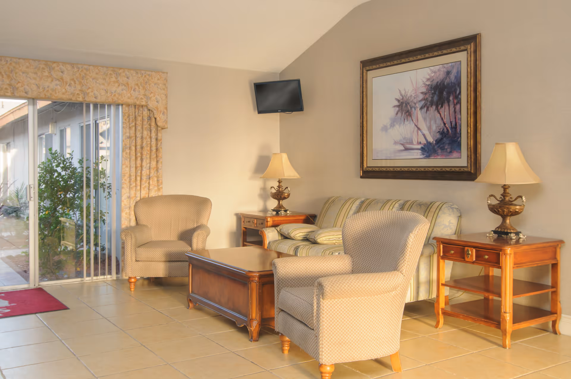 A cozy living room area with two beige armchairs, a striped sofa, a wooden coffee table, and two wooden side tables with lamps. A framed painting of a sailboat and palm trees hangs on the wall above the sofa. There is a small wall-mounted TV in the corner and a sliding glass door with floral curtains leading outside.