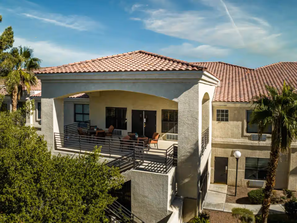 Two-story Mediterranean-style exterior showing a covered second-floor balcony with tables and chairs, terracotta tile roof and surrounding palm trees.
