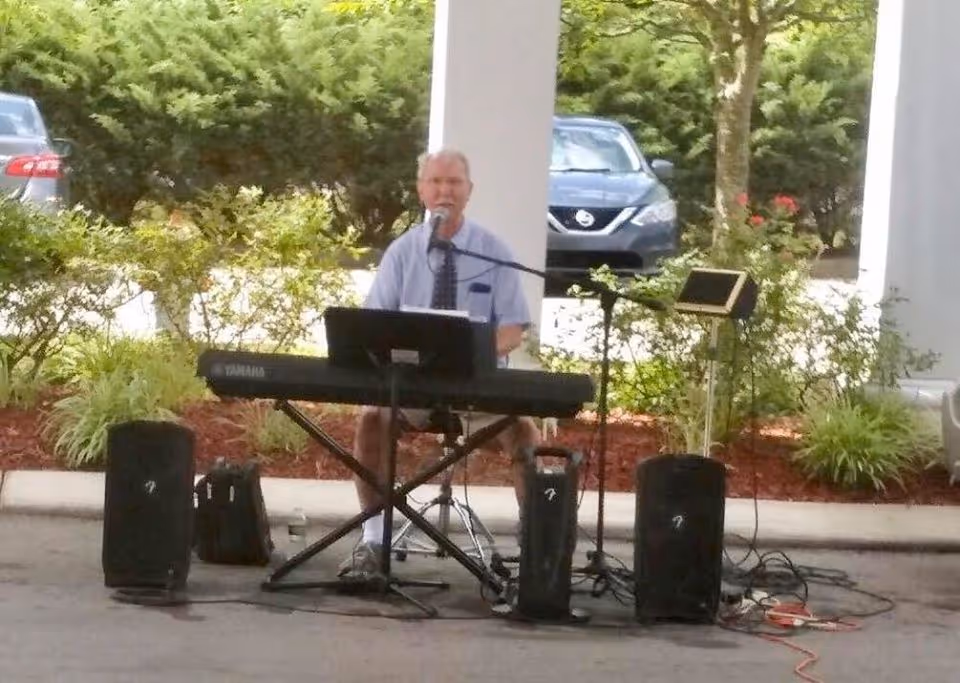 An elderly man is playing a Yamaha keyboard and singing into a microphone set up on a stand. He is seated outdoors under a covered area with plants and parked cars visible in the background. There are speakers and audio equipment around him.