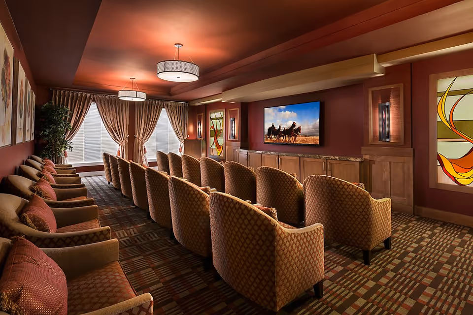 A cozy theater-style lounge with rows of upholstered armchairs facing a wall-mounted television in a warmly decorated room.