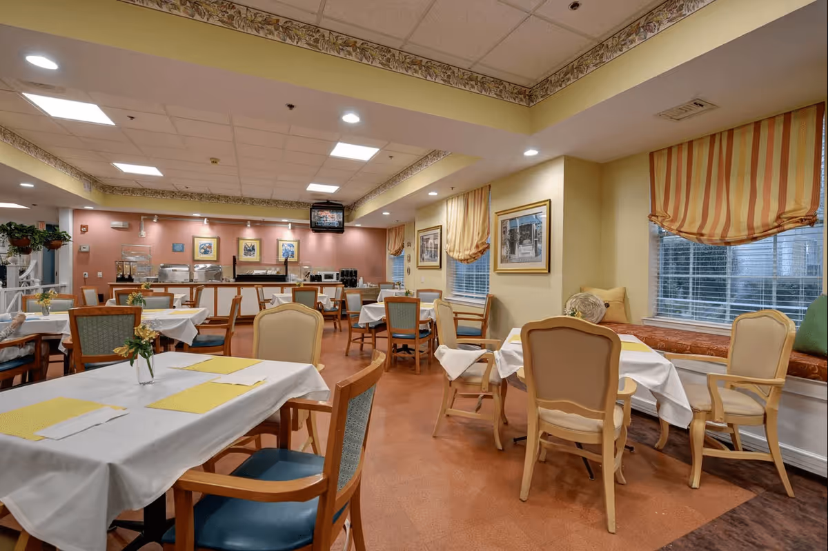 A dining room in Ville Ste Marie Senior Living with several tables covered in white tablecloths and yellow placemats. The room features wooden chairs with blue and beige cushions, large windows with striped yellow and orange curtains, framed pictures on the walls, and a buffet area with food warmers and a television mounted above it.