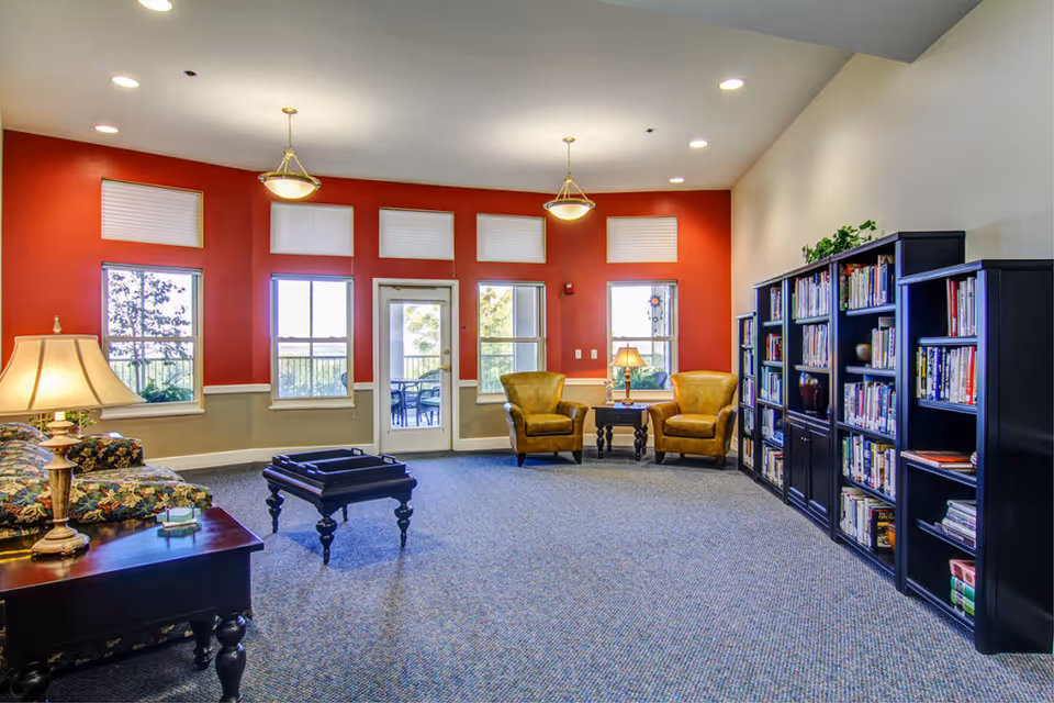 A bright and spacious senior living community lounge with red and beige walls, large windows, and a glass door leading outside. The room features two yellow armchairs with a small table and lamp between them, a floral-patterned sofa, a dark wooden coffee table, and several black bookshelves filled with books. Ceiling lights and table lamps provide warm lighting.