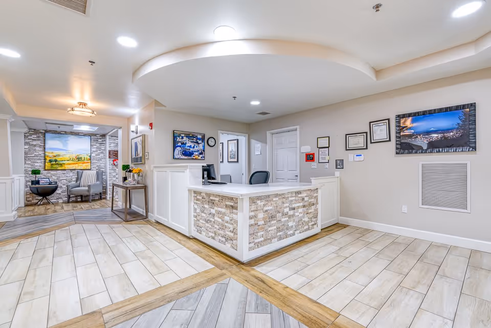 Reception area of Sellwood Senior Living featuring a central desk with a computer and chair, light-colored tiled flooring with wood accents, framed pictures and certificates on the walls, and a seating area with chairs and a table in the background.