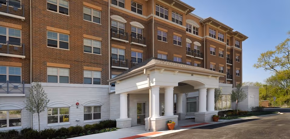 Exterior view of a multi-story senior living facility building with a brick facade and white lower walls. The entrance features a covered portico with white columns and a small driveway. There are small trees and shrubs planted along the building, and the sky is clear and blue.
