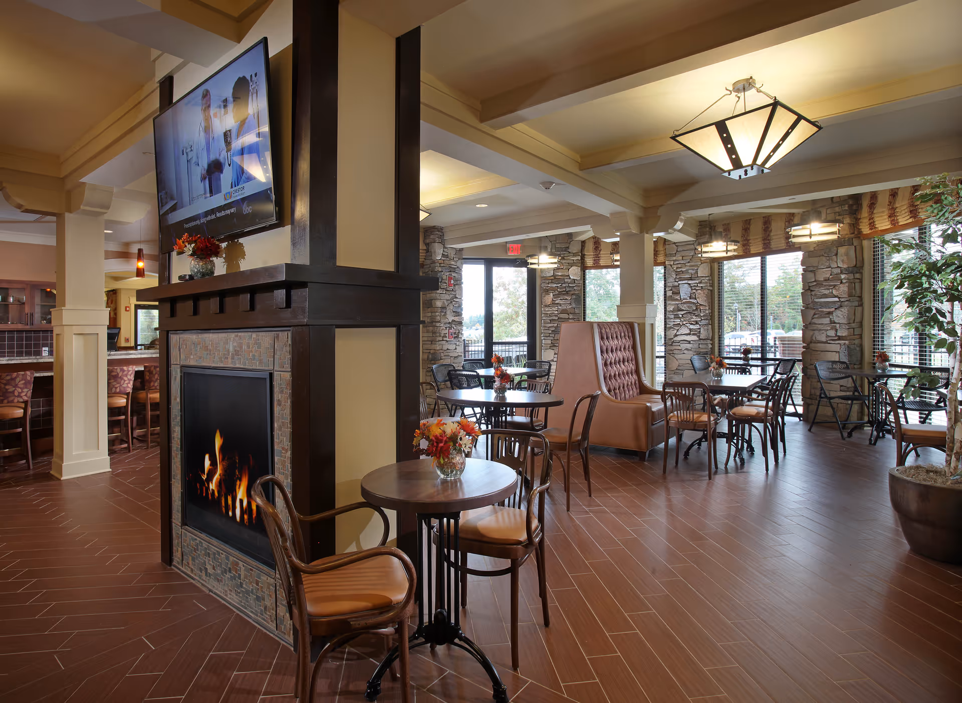 A cozy dining area with multiple tables and chairs arranged around a central fireplace. The fireplace has a mounted flat-screen TV above it. The room features large windows with stone walls, letting in natural light. There are decorative flowers on the tables and a large potted plant in the corner. The floor is covered with brown tiles, and the ceiling has exposed beams with hanging light fixtures.