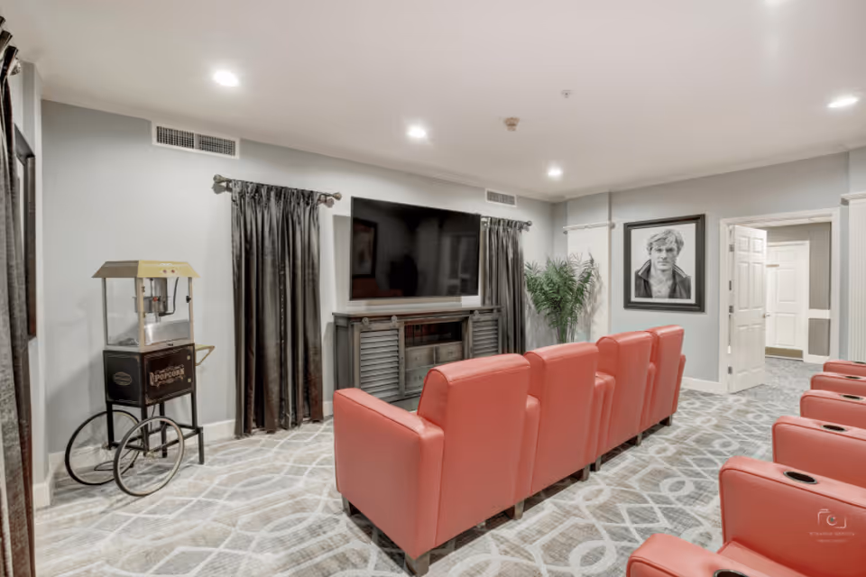 A cozy media room with two rows of red theater-style chairs facing a large flat-screen TV mounted on a gray wall. There is a popcorn machine on a cart to the left, dark curtains on the windows, a framed black and white portrait on the right wall, and a potted plant near the corner. The room has patterned carpet flooring and recessed ceiling lights.