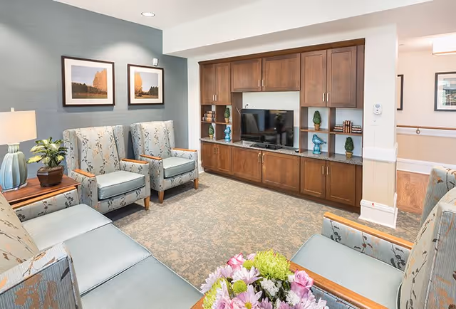 A cozy senior living common area with light blue patterned armchairs and a matching sofa arranged around a wooden entertainment center with a flat-screen TV. The room has soft carpeting, a side table with a lamp and plant, framed landscape pictures on the wall, and a bouquet of flowers in the foreground.