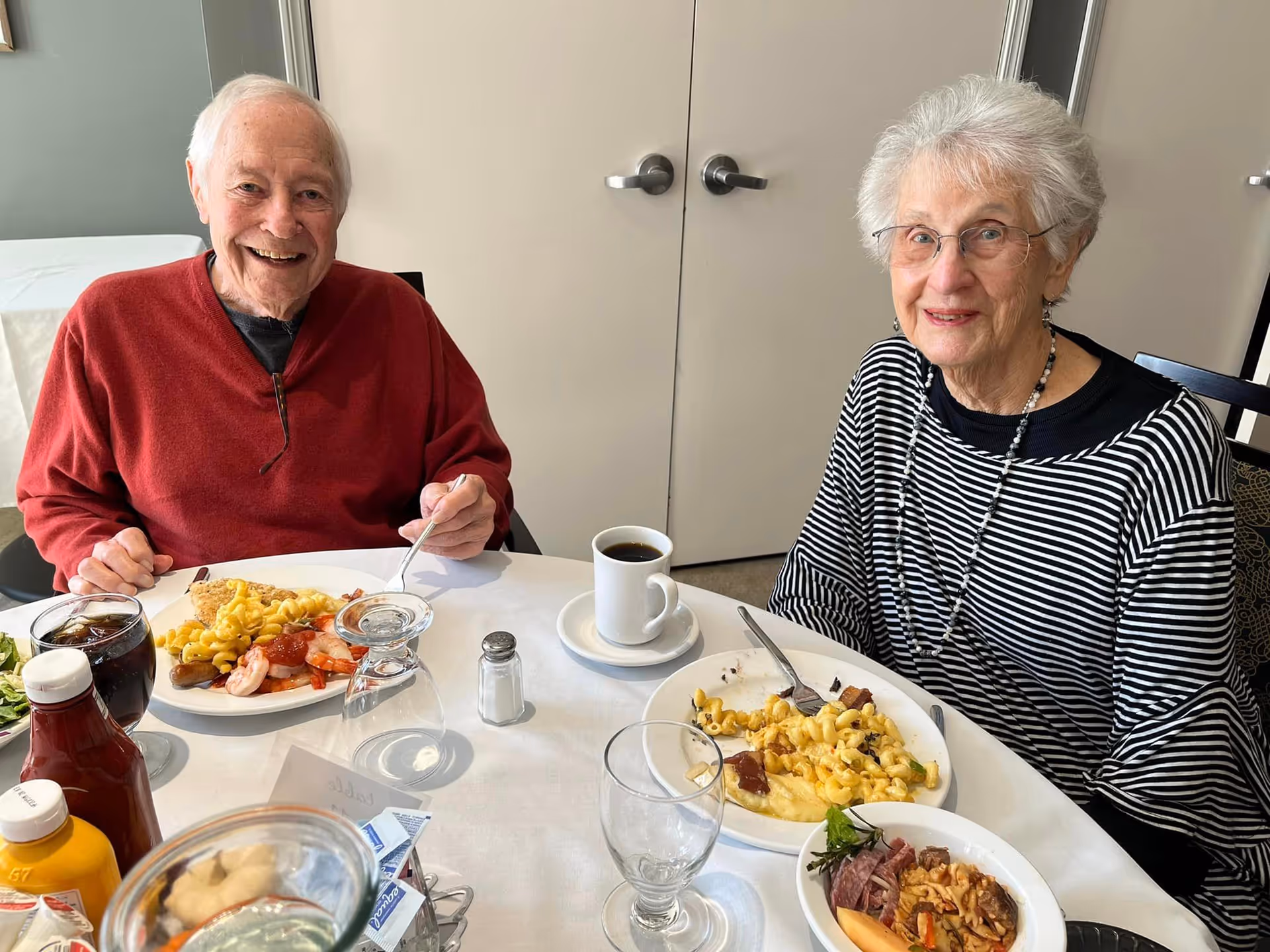 An elderly man and woman sitting at a round dining table with plates of food in front of them. The man is wearing a red sweater and smiling, while the woman is wearing glasses and a black and white striped top, also smiling. The table has various food items, drinks, and condiments on it, with a plain wall and double doors in the background.
