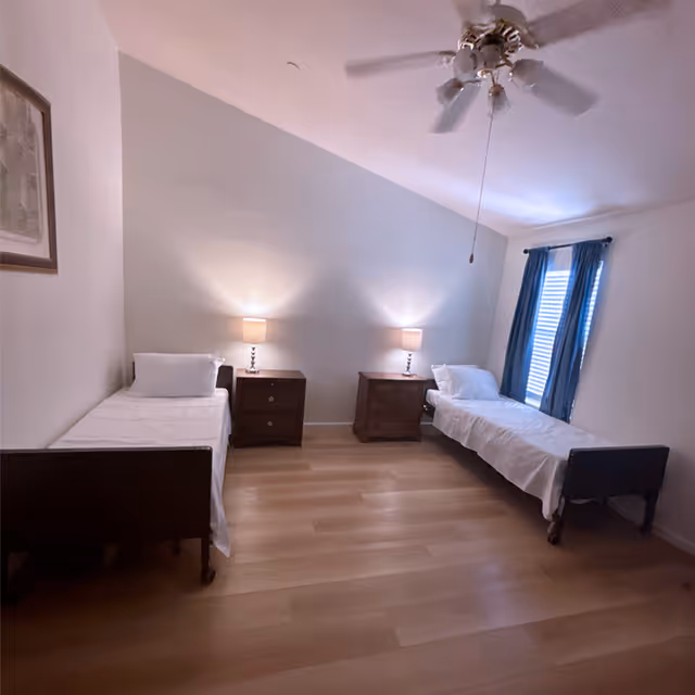 A simple bedroom with two single beds placed against opposite walls. Each bed has white bedding and a pillow. Between the beds are two wooden nightstands, each with a lit table lamp. A window with blue curtains is on the right wall, and a ceiling fan with lights hangs from the ceiling. The floor is wooden, and a framed picture is hung on the left wall.