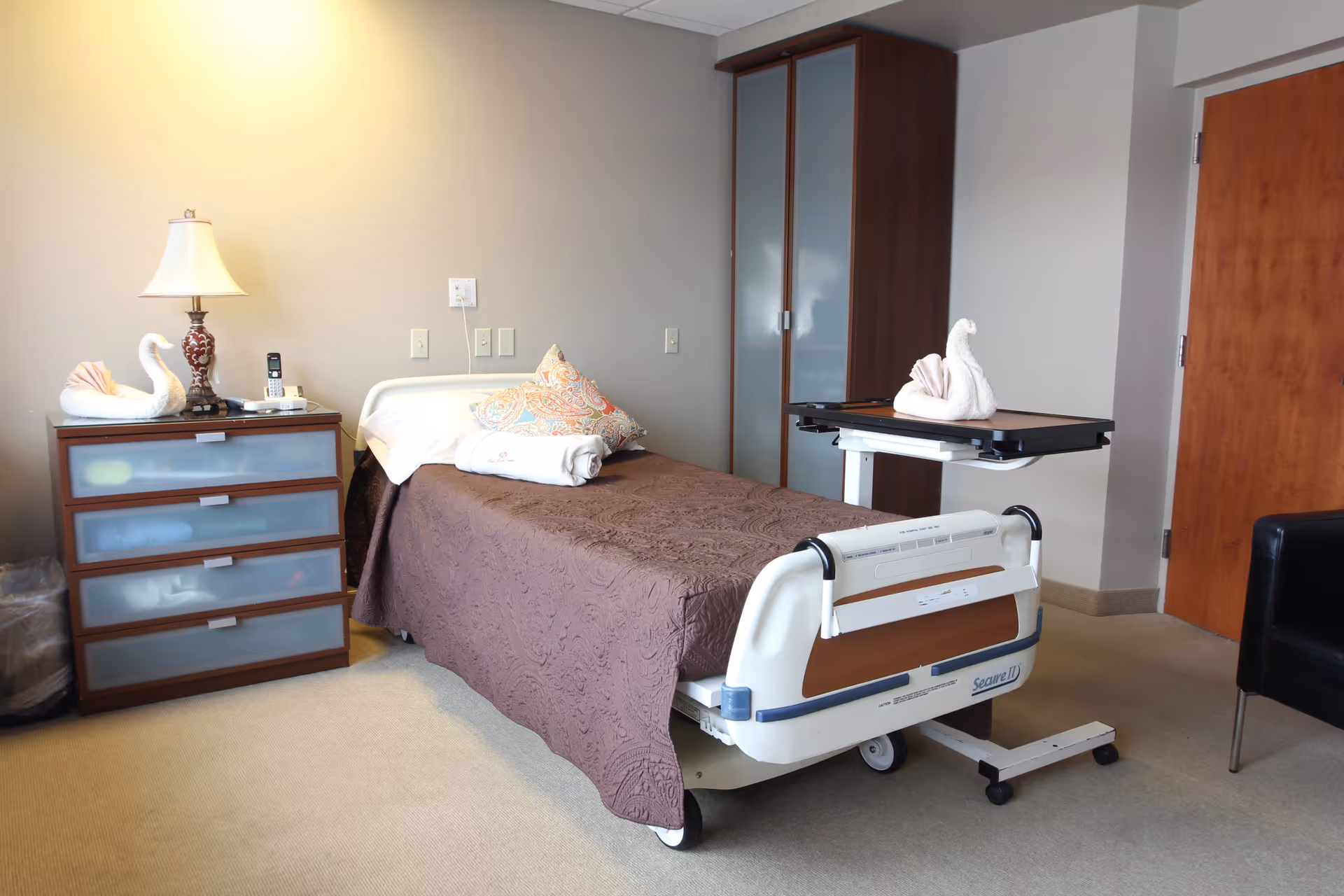 A furnished patient bedroom with a single hospital bed, bedside table and lamp, wardrobe, and overbed table.