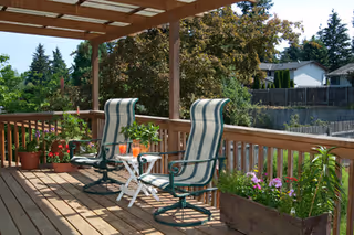 A wooden outdoor deck with two striped cushioned chairs and a small white table between them holding two glasses of orange drink. The deck is surrounded by wooden railings and has potted plants and flowers along the edges. Trees and neighboring houses are visible in the background under a clear sky.