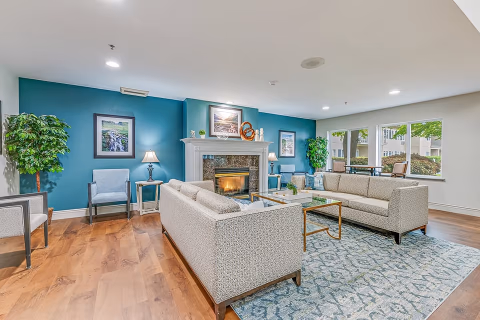 A bright and spacious living room in a senior living facility featuring two patterned sofas arranged around a glass coffee table on a blue patterned rug. The room has a fireplace with a white mantel, flanked by two table lamps and framed artwork on a teal accent wall. There are also two armchairs, potted plants, and large windows letting in natural light with a view of outdoor greenery and patio seating.