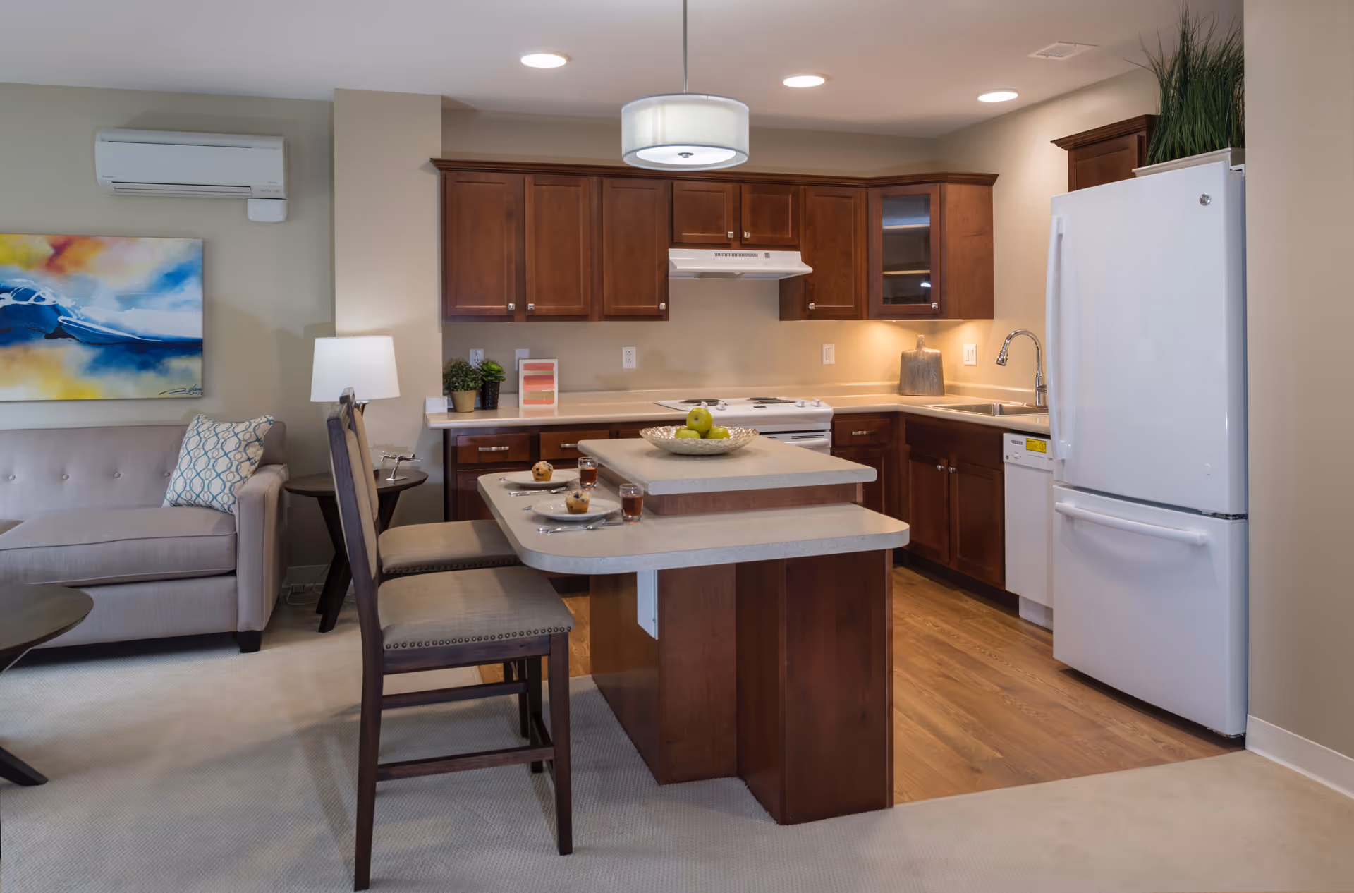 A modern kitchen area with wooden cabinets, a white refrigerator, a stove with a hood, a dishwasher, and a double sink. There is a kitchen island with two chairs, set with plates, utensils, muffins, and glasses of juice. To the left, part of a beige sofa with a patterned pillow and a side table with a lamp are visible. A colorful abstract painting hangs on the wall above the sofa.