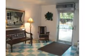A small waiting area with a wooden bench and chair, a floor lamp, a framed picture on the wall, a plant hanging on the wall, and a glass door leading outside. The floor has a checkered pattern with blue and white tiles.