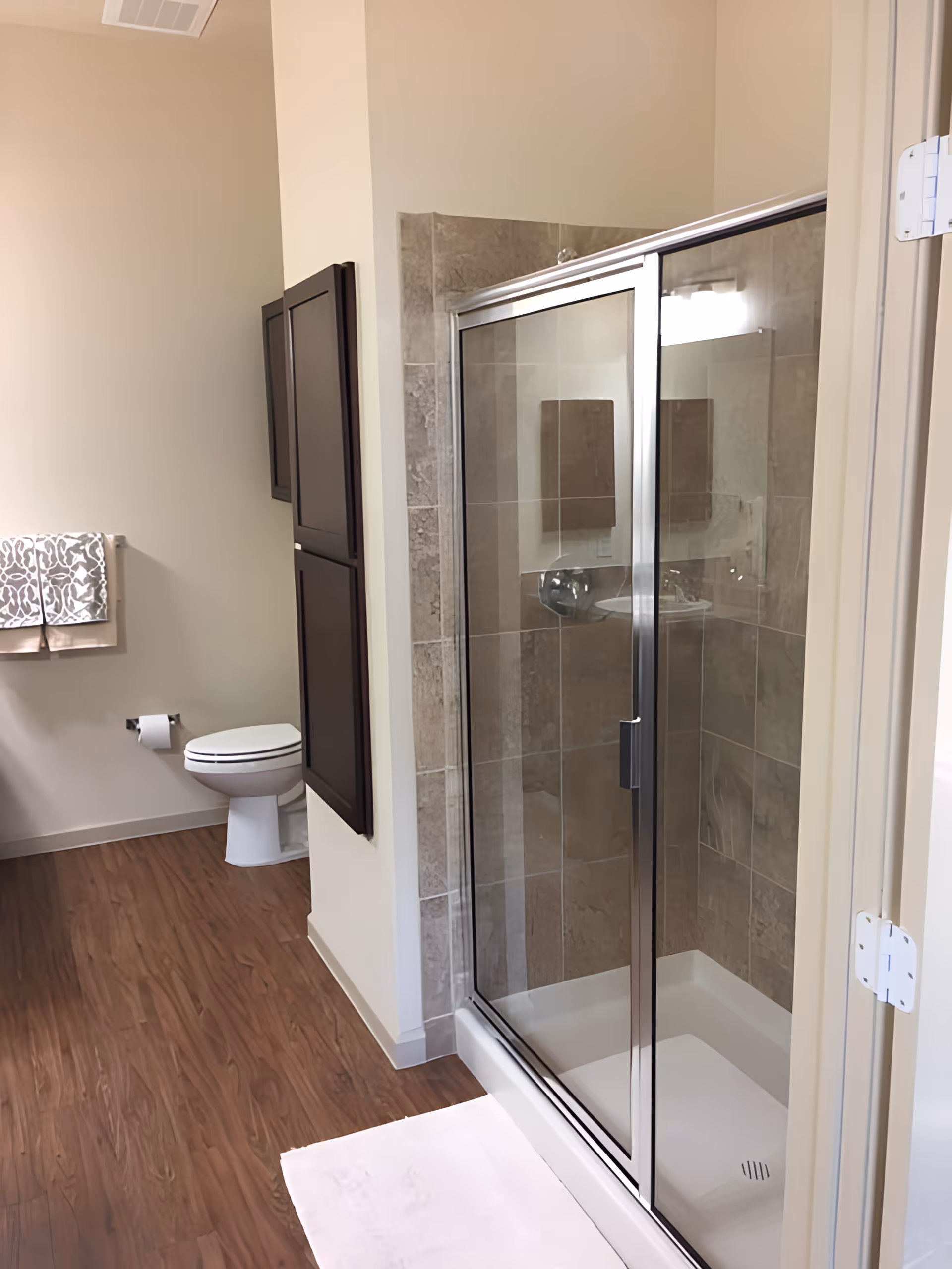 A bathroom with a glass-enclosed shower featuring beige tiles, a white toilet, a wall-mounted dark wood cabinet, a towel rack with two patterned towels, and wood-look flooring.