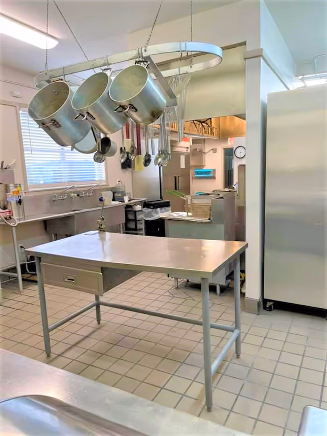 A clean commercial kitchen with a stainless steel work table in the center, large pots and utensils hanging from a ceiling rack, a sink under a window with blinds, and various kitchen appliances and storage units in the background.