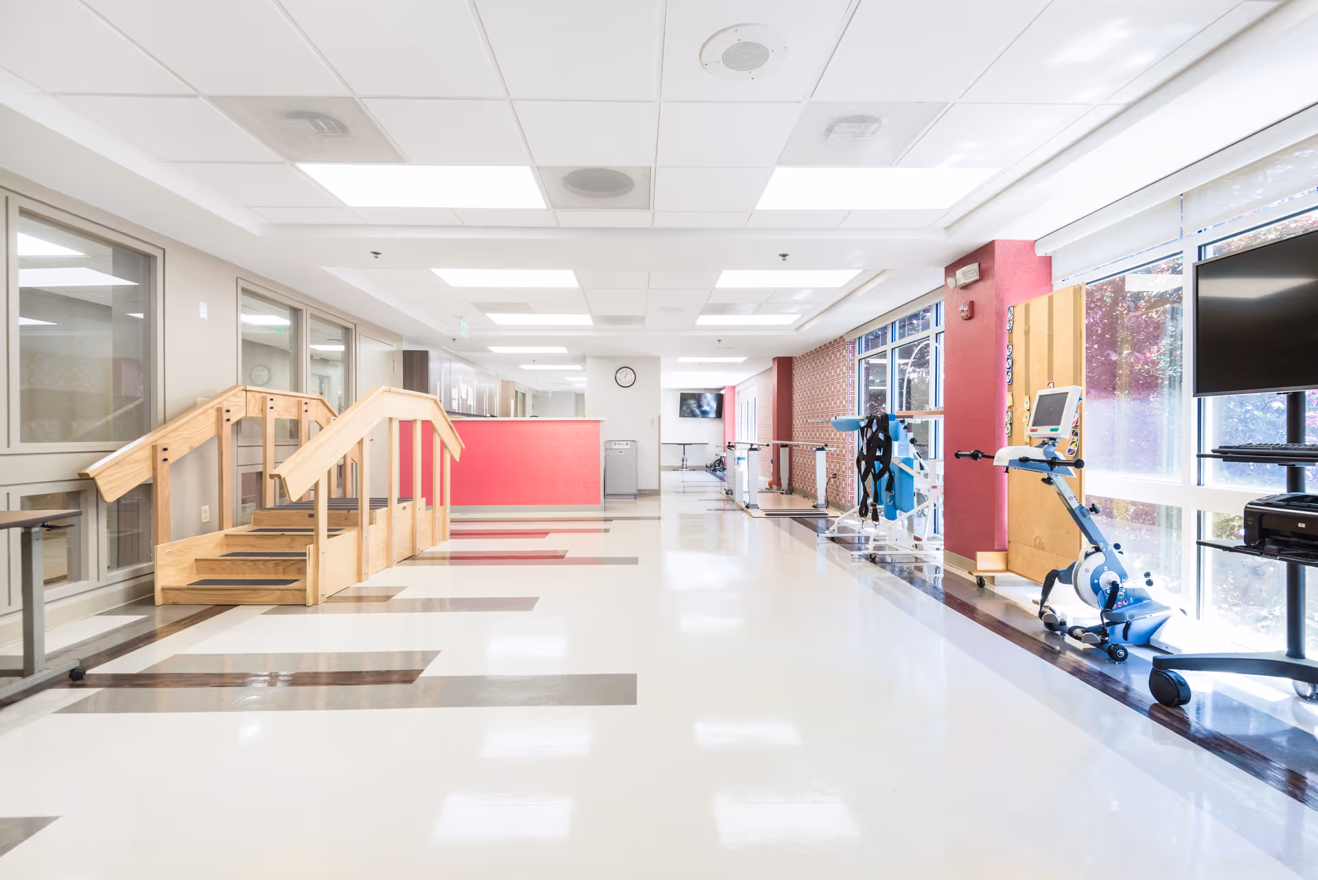 Bright rehabilitation therapy corridor with parallel bars, exercise machines, and wooden practice stairs.