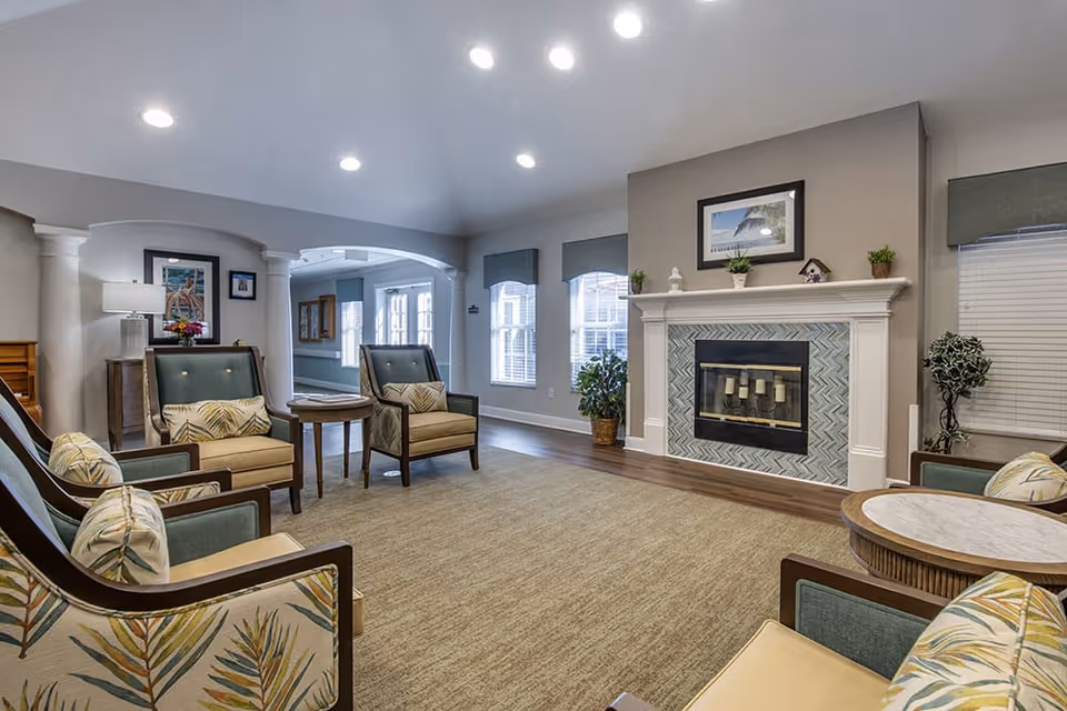 Bright, furnished common room with armchairs and side tables arranged around a fireplace under recessed lighting.
