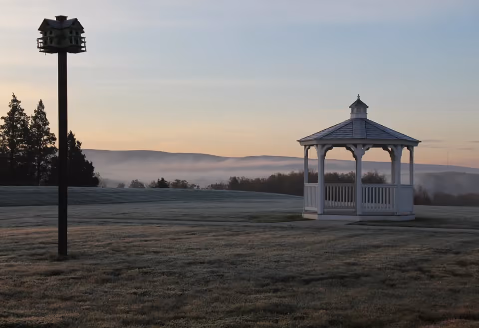 A serene outdoor scene at dawn featuring a white wooden gazebo on a grassy field with a birdhouse mounted on a tall pole to the left. In the background, there are trees and rolling hills partially covered by morning mist under a clear sky.