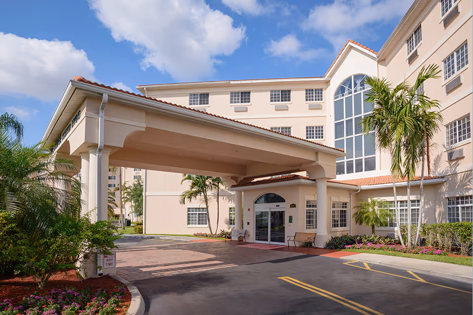 Front entrance of a beige senior living building with a covered porte-cochere, palm trees, and large windows.