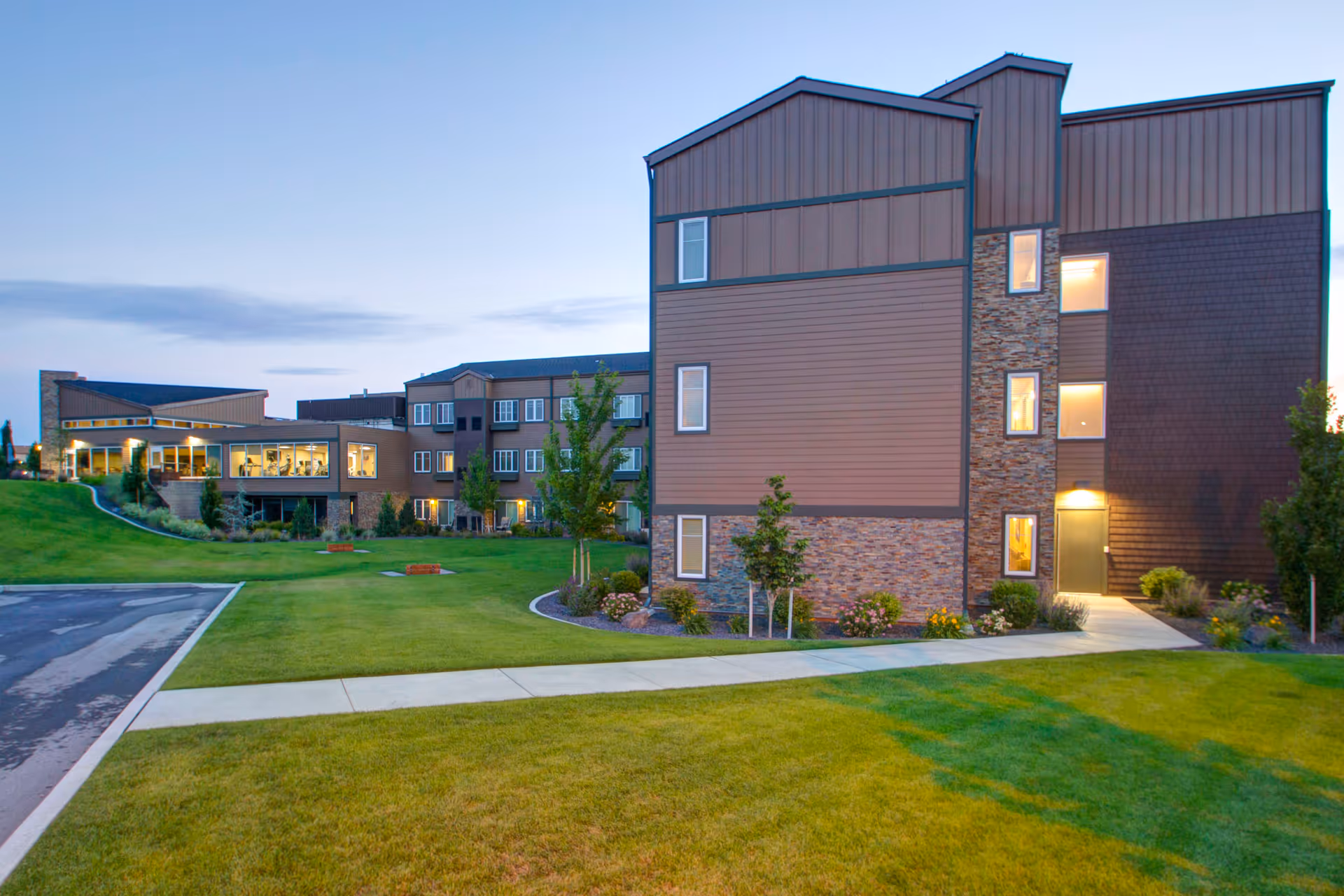 Exterior view of a multi-story senior living facility building with brown siding and stone accents, surrounded by a well-maintained lawn and small trees, under a clear evening sky.