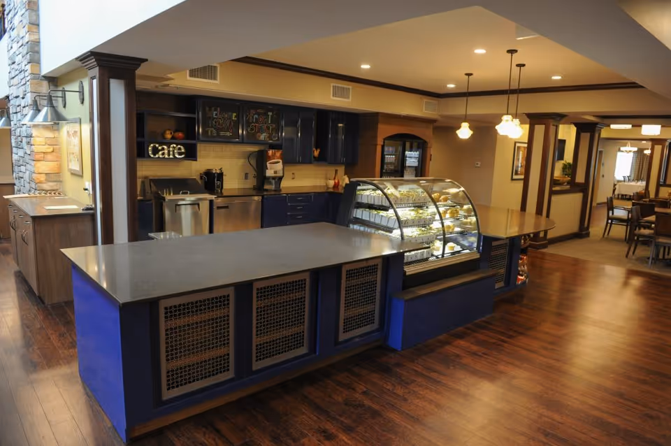 Interior view of a senior living facility cafe area with a large counter, a glass display case filled with pastries, dark blue cabinetry, and warm lighting. The space features wooden flooring and a dining area visible in the background.